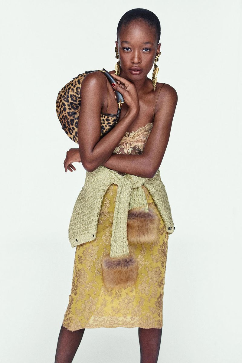 Model in a lace slip dress with a green sweater tied around the waist holding a leopard print handbag against a white background