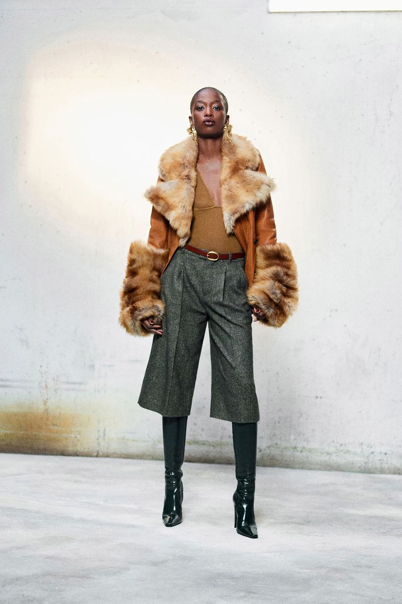 Fashionable woman in a tan faux fur coat and green culottes poses in front of a textured white wall in a studio setting.