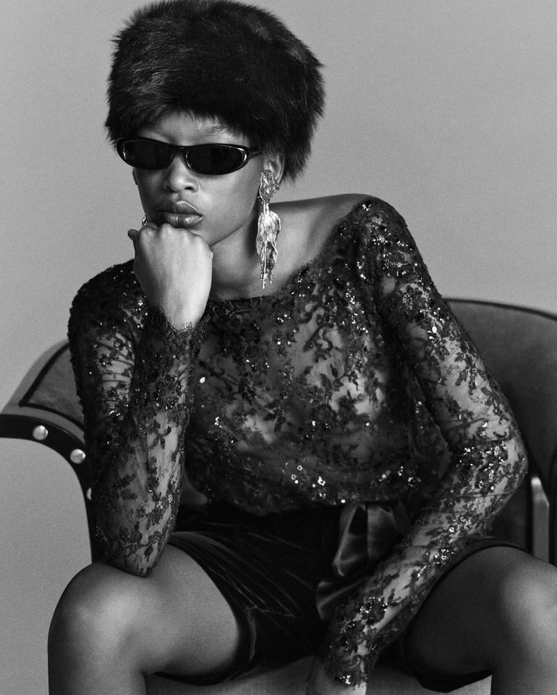 Model wearing dark sunglasses, a furry hat, and a black lace top seated indoors against a plain background