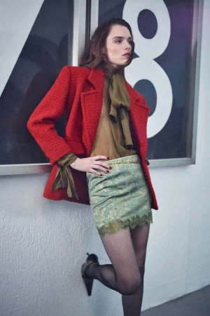 Model wearing a red textured blazer, olive blouse, and patterned skirt leaning against a white textured wall