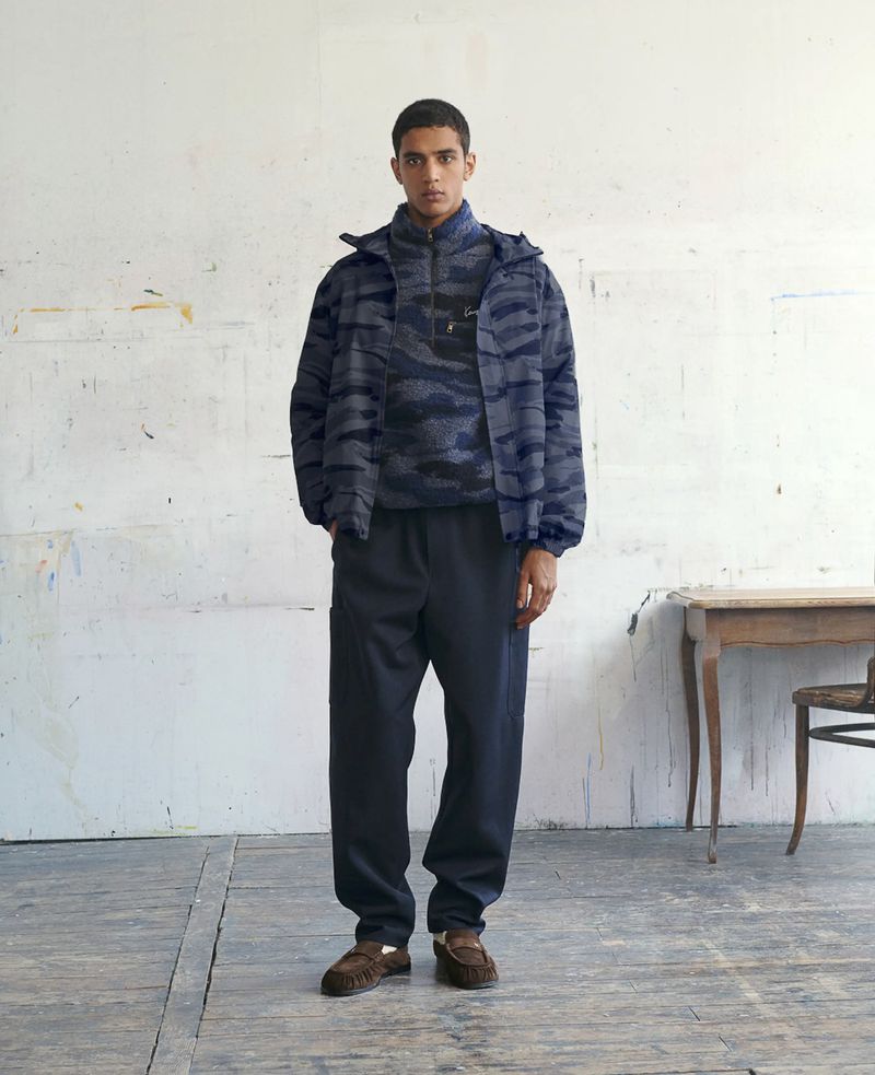 Male model wearing a blue patterned fleece jacket and dark trousers standing in an artist studio.