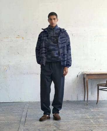 Male model wearing a blue patterned fleece jacket and dark trousers standing in an artist studio.