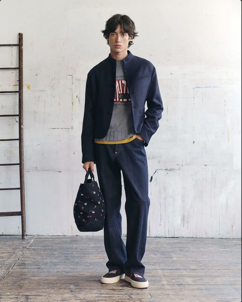 Young male model wearing navy jacket and jeans holding patterned tote bag inside studio