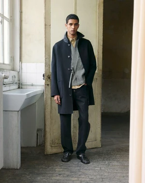 A young male model standing indoors wearing a dark coat, grey vest, and trousers, photographed for Kenzo.