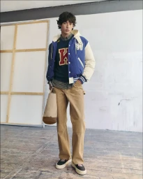 Model wearing a blue varsity jacket, sweater, and brown trousers holding a tote bag in a studio setting for Kenzo.