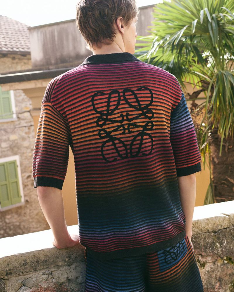 A man viewed from behind wearing a striped knit polo shirt and matching shorts against a rustic stone building and tropical foliage background.