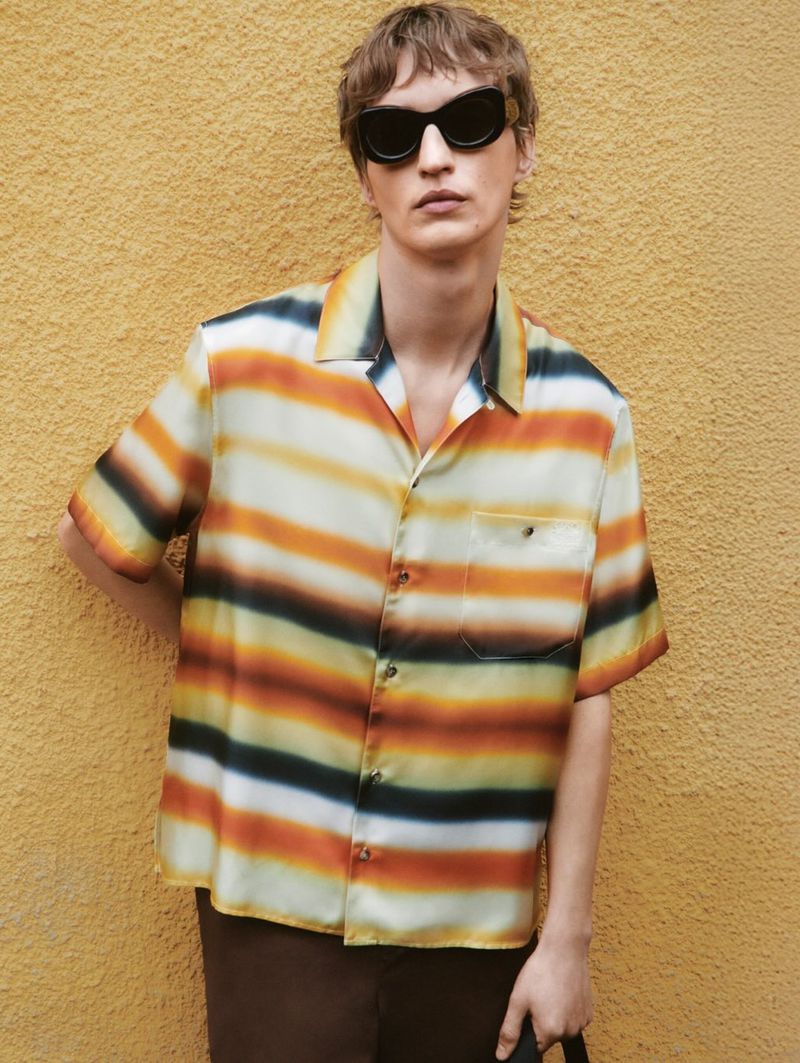 A fair-skinned adult male wearing a striped camp shirt and sunglasses stands against a textured yellow wall in an eye-level outdoor shot.