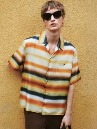 A fair-skinned adult male wearing a striped camp shirt and sunglasses stands against a textured yellow wall in an eye-level outdoor shot.