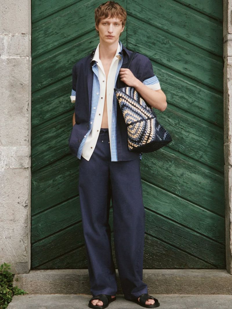 Male model in layered blue and white shirt holding a patterned tote bag in front of a green wooden door