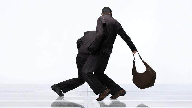 Two people in matching brown outfits walking on a glossy white surface, one holding a brown handbag. Simple studio setting, fashion focused.