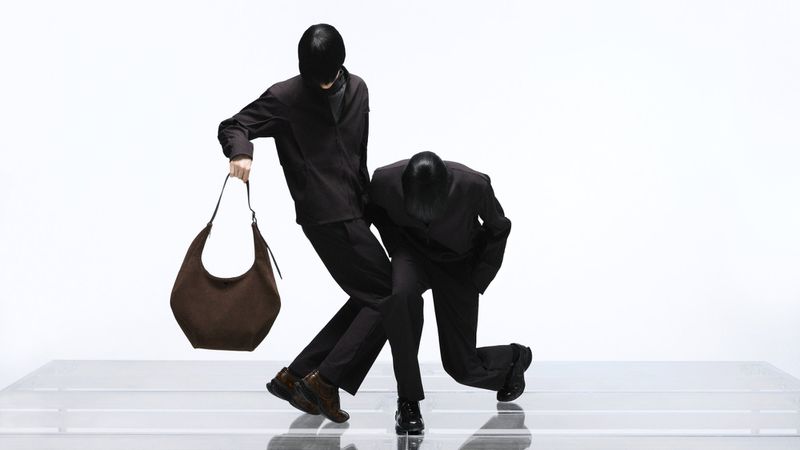 Two figures in dark clothing posing dynamically against a bright white background, one holding a brown shoulder bag
