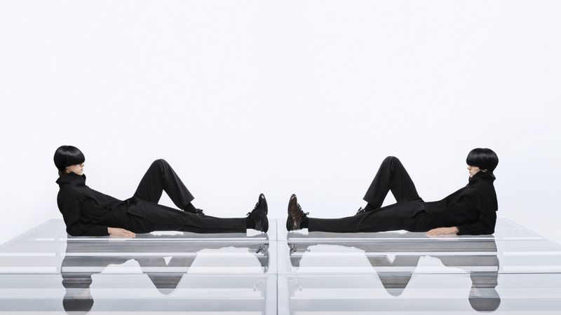 Two models in black outfits lying symmetrically on glass platforms against a plain white background for Puma.