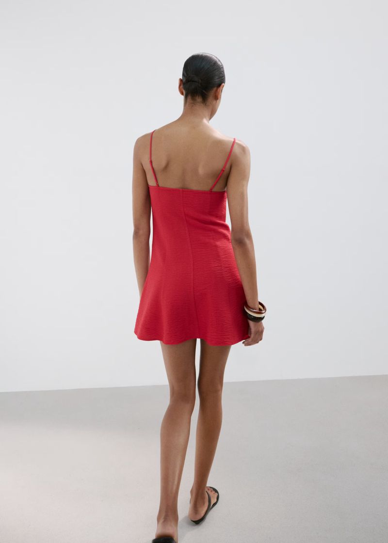 Model standing facing away wearing a short red slip dress against a plain white studio background