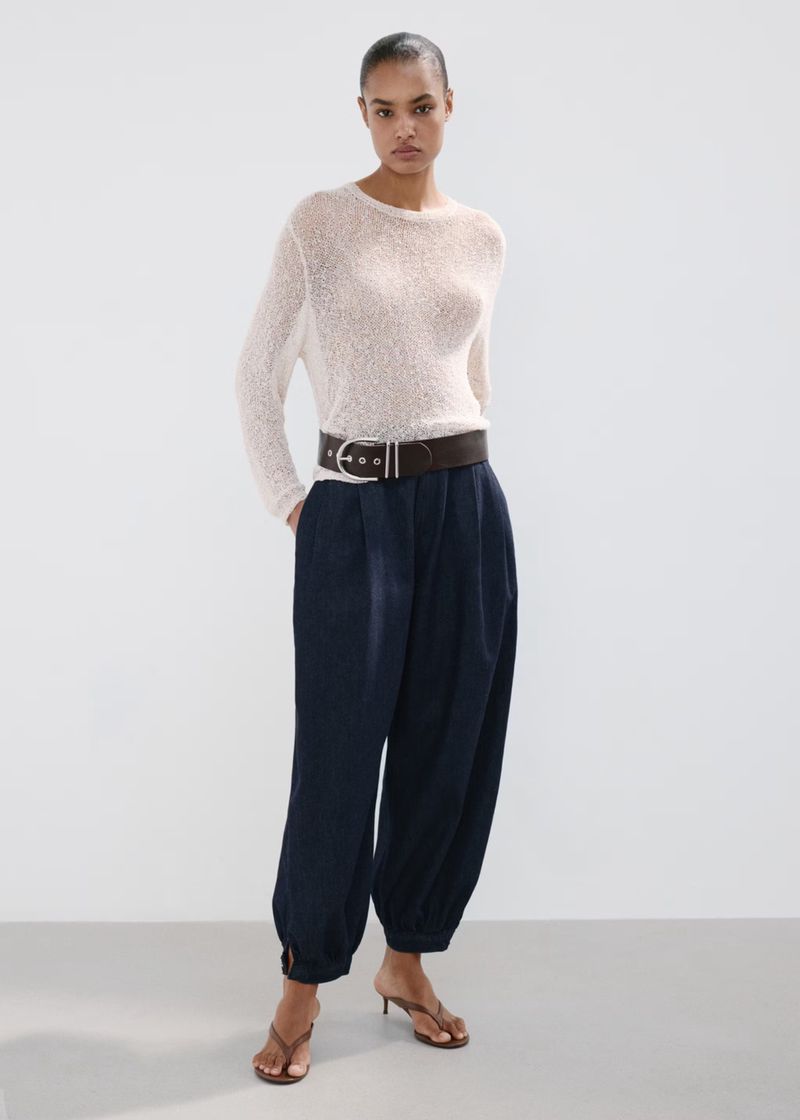 Model Ana Beatriz Cortes wearing a white textured top, dark trousers, and a wide belt against a neutral studio backdrop.