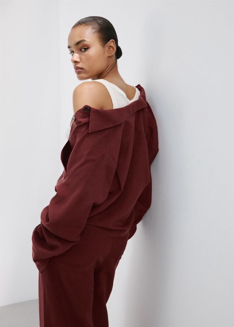 Model wearing a burgundy jacket and matching trousers leaning against a white studio wall