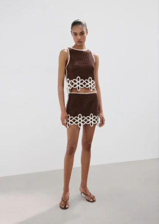 Model standing on a grey floor against a white wall wearing a brown knit two-piece outfit with white flower cutouts.