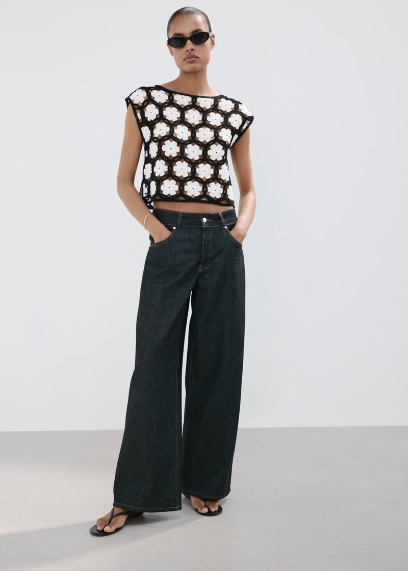 Model wearing black sunglasses, black and white patterned top, and dark wide-leg jeans against white seamless background.