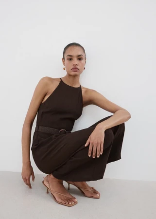 Model in a dark brown halterneck jumpsuit crouching against a plain white studio background.