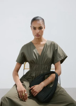 Model seated wearing an olive green midi dress holding a black woven handbag against a white background