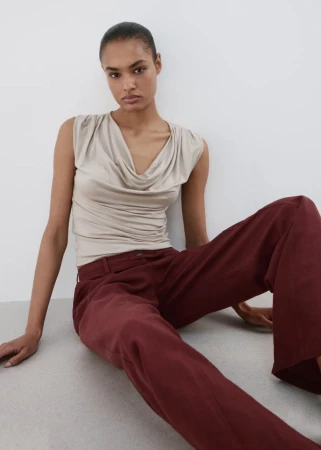Model seated on the floor wearing a draped neutral top and burgundy trousers against a clean white background