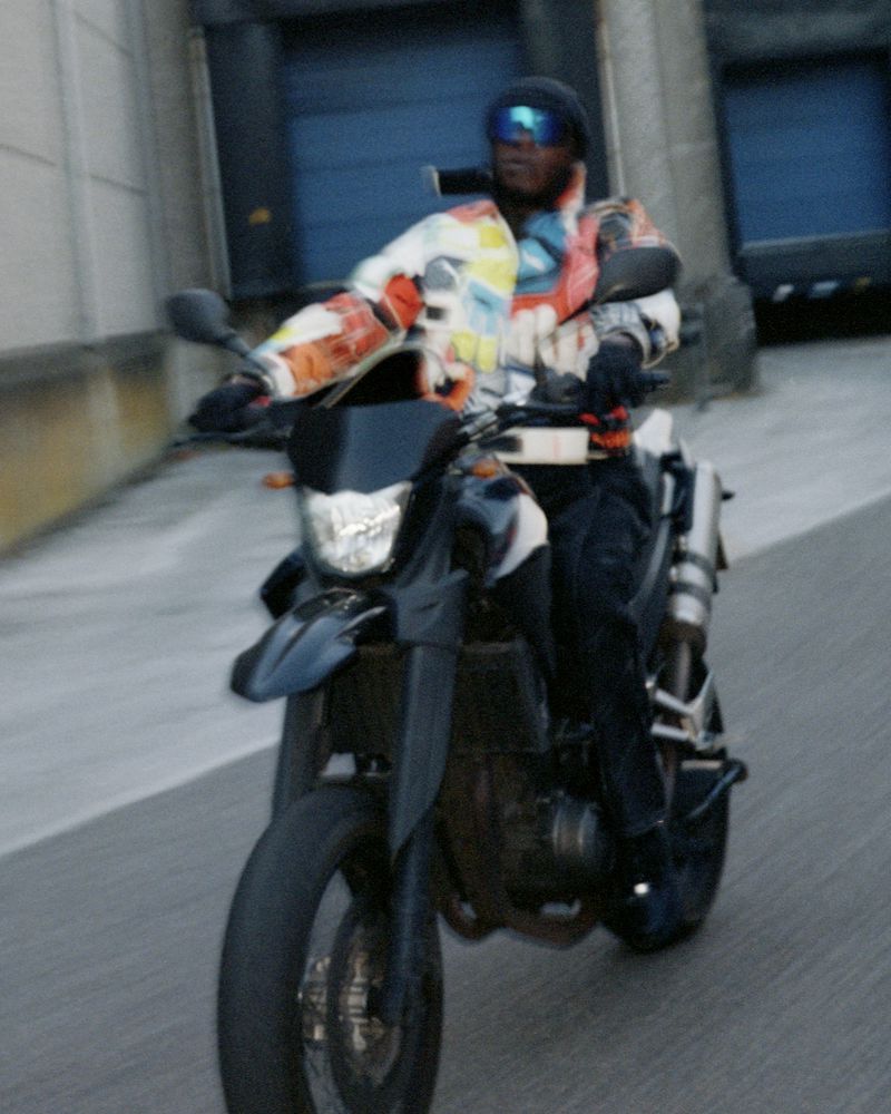 Man rides a black motorbike on an urban road. The rider wears goggles and a patterned jacket, creating a dynamic street scene.