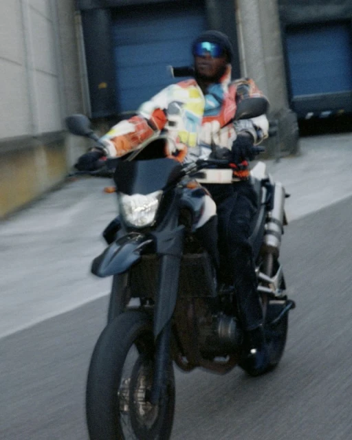 Man rides a black motorbike on an urban road. The rider wears goggles and a patterned jacket, creating a dynamic street scene.