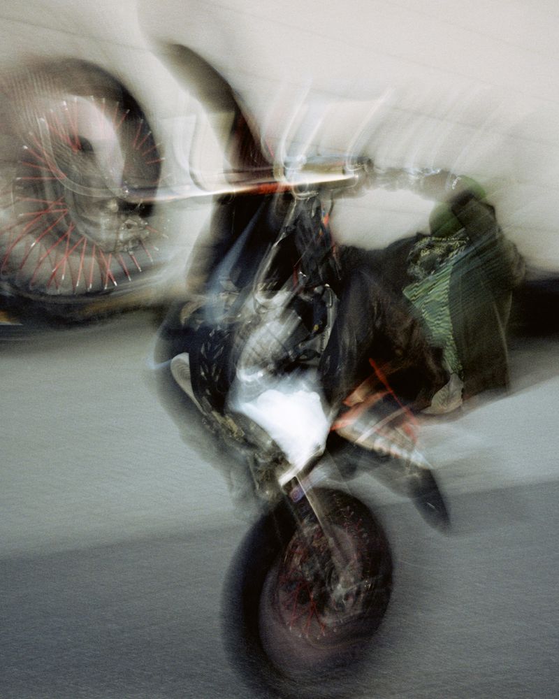 Abstract long exposure of a motorcycle showing motion blur, red wheels and indistinct shapes against a pale, neutral background.