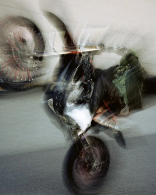 Abstract long exposure of a motorcycle showing motion blur, red wheels and indistinct shapes against a pale, neutral background.