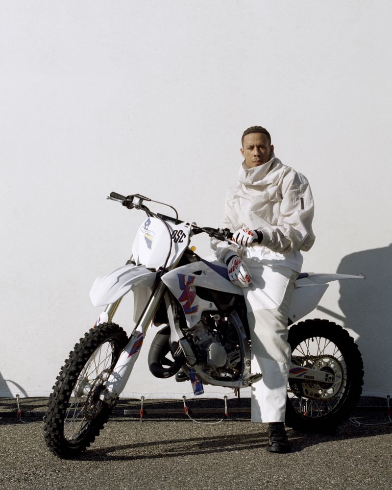 A man in a white racing suit poses with a white dirt bike against a light grey wall on an asphalt surface in an outdoor shot.