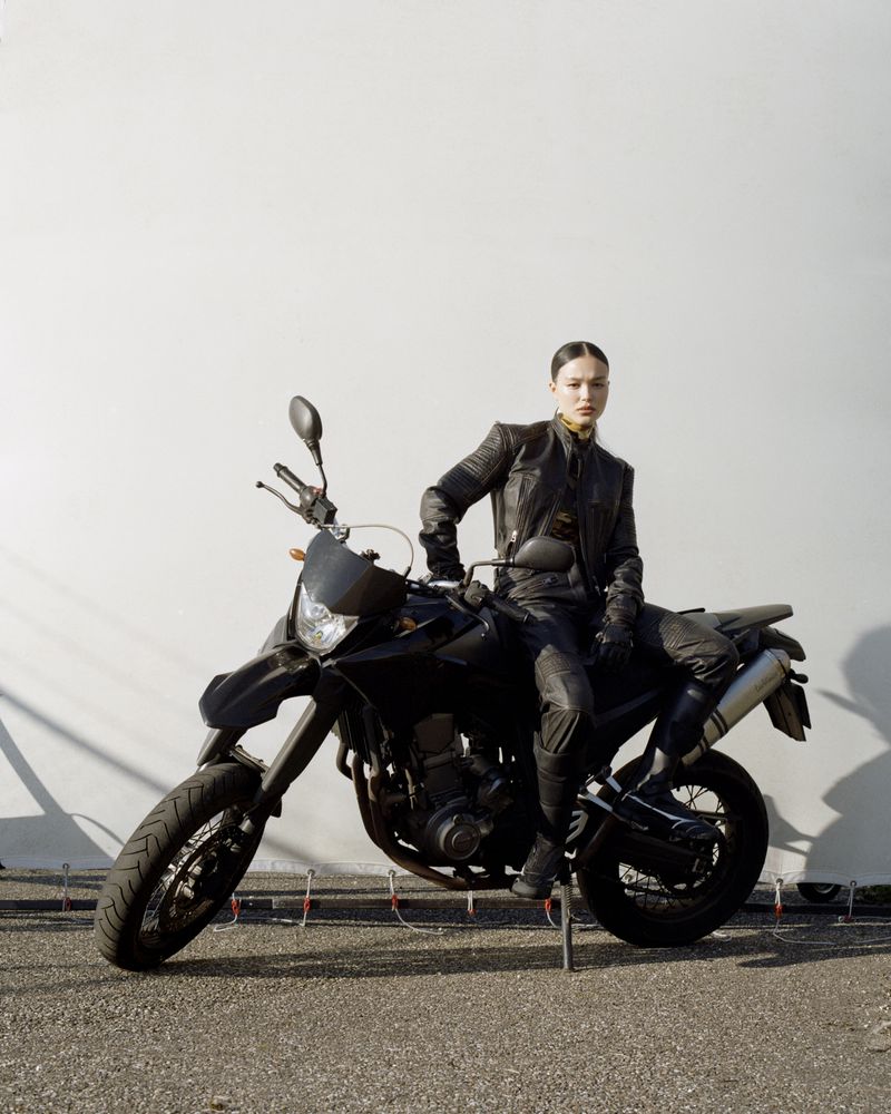 Adult female poses on a black motorcycle outdoors wearing a leather jacket. Neutral expression, full-body shot against a white wall.
