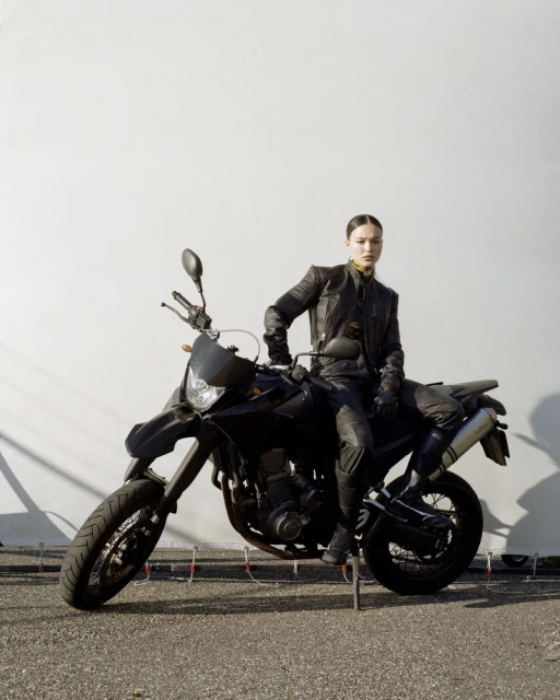 Adult female poses on a black motorcycle outdoors wearing a leather jacket. Neutral expression, full-body shot against a white wall.
