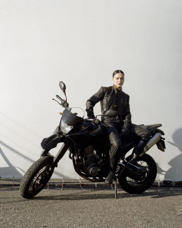 Adult female poses on a black motorcycle outdoors wearing a leather jacket. Neutral expression, full-body shot against a white wall.