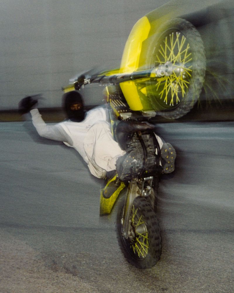 Adult in a white outfit and black balaclava performs a wheelie stunt on a yellow and grey dirt bike on an asphalt surface.