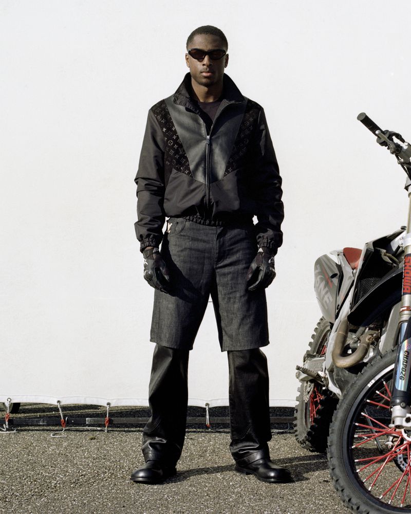Fashionable Black man in jacket and shorts poses with dirt bike against a white backdrop, wearing sunglasses, gloves, and boots.
