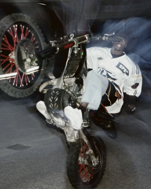Man performing wheelie on dirt bike. Adult male wearing a jersey and jeans on a motorcycle with red spokes on the wheels. 