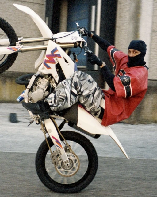 A motorcycle stunt shows a person in a black beanie doing a wheelie on a white, blue, and pink motorcycle, with a building background.