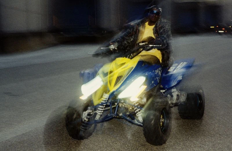 Adult male driving a yellow and blue all-terrain vehicle on a street, wearing a black leather jacket, black sunglasses, and a hat.