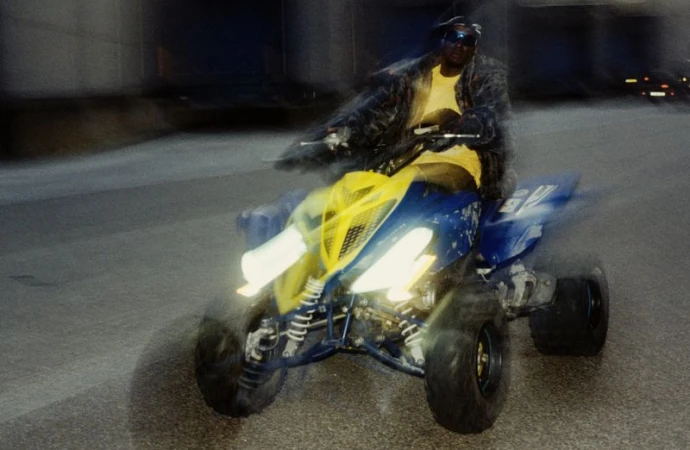 Adult male driving a yellow and blue all-terrain vehicle on a street, wearing a black leather jacket, black sunglasses, and a hat.