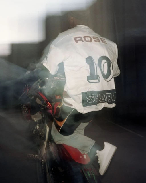 Person on motorcycle with shirt displaying 'Rose' and '10' riding on an outdside street with motion blur and poor lighting