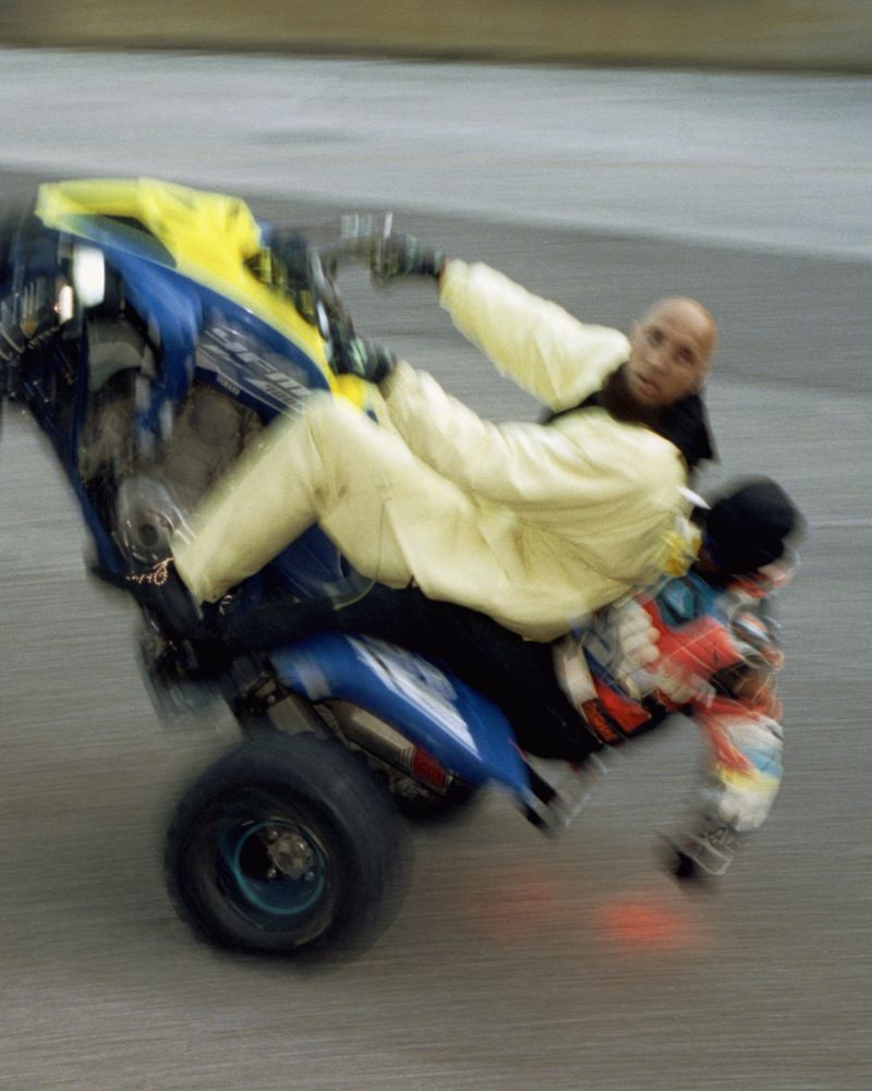 Two people on an ATV performing a wheelie stunt with motion blur, wearing casual attire and protective gloves on a paved surface.