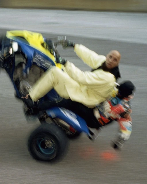 Two people on an ATV performing a wheelie stunt with motion blur, wearing casual attire and protective gloves on a paved surface.