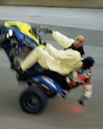 Two people on an ATV performing a wheelie stunt with motion blur, wearing casual attire and protective gloves on a paved surface.