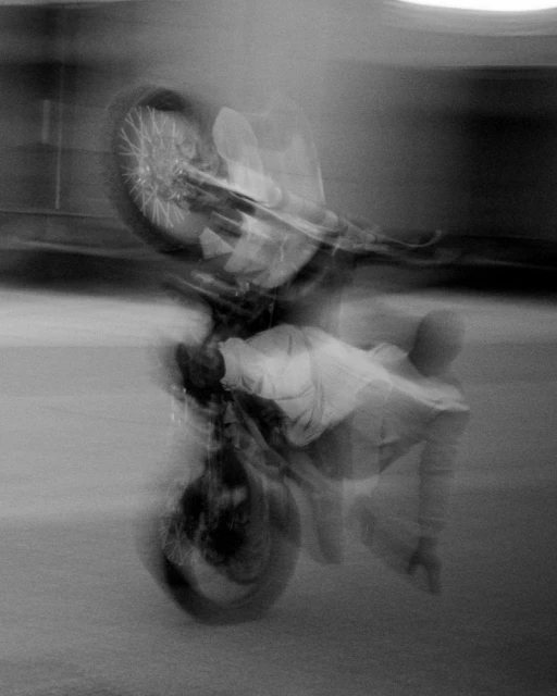 Monochrome photo of a man riding a motorcycle in motion blur, performing a wheelie on a paved surface with blurred background.