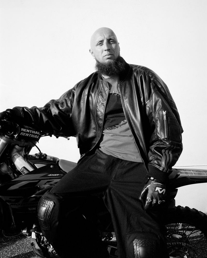 Black and white shot of a bald adult male with a beard, wearing a leather jacket, seated on a motorcycle against a white backdrop.