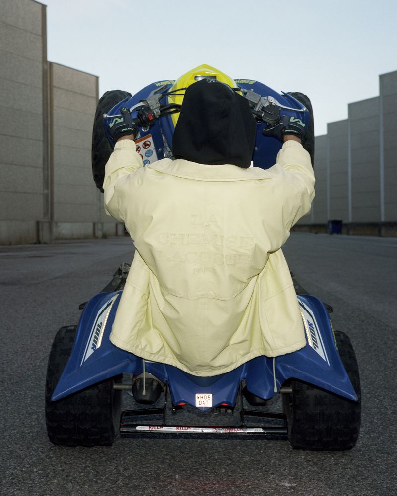 Person in yellow jacket on a blue ATV performing a wheelie on an asphalt surface against a concrete wall background.