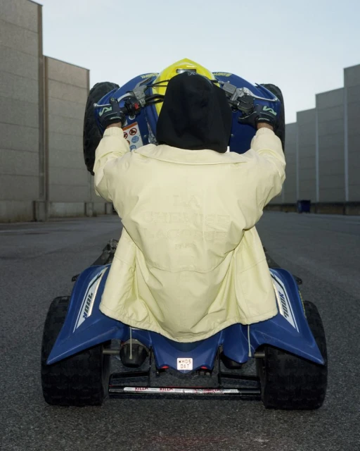 Person in yellow jacket on a blue ATV performing a wheelie on an asphalt surface against a concrete wall background.