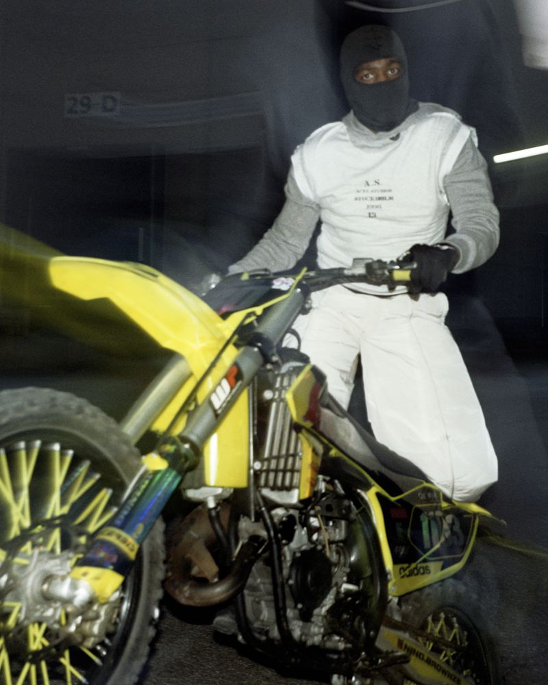 Man in racing suit and balaclava sitting on a yellow and black dirt bike with a blurred background in a dark garage or warehouse.