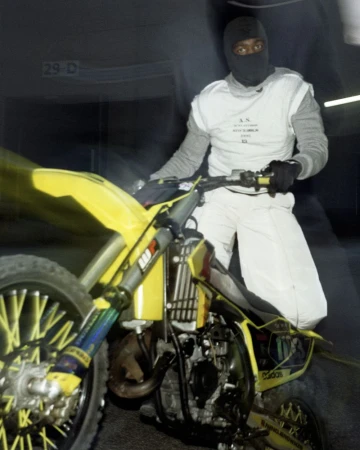 Man in racing suit and balaclava sitting on a yellow and black dirt bike with a blurred background in a dark garage or warehouse.