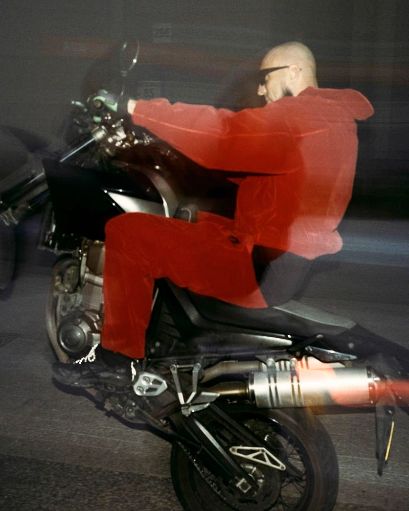 A man in a red hooded tracksuit does a motorcycle wheelie at night with motion blur and direct flash.