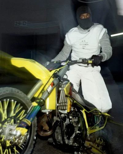 A Black man wearing a black balaclava and white apparel seated on a yellow dirt bike in an underground garage.
